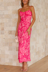 A woman smiles and looks towards the camera, wearing a long, pink floral print dress with spaghetti straps and a ruched bodice.