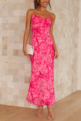 A woman stands with a white clutch in her hand, wearing a long, pink floral print dress with spaghetti straps and a ruched bodice.