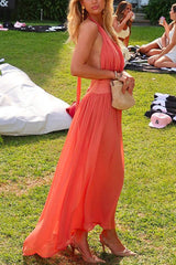 Woman wearing a coral halter neck maxi dress with a flowy skirt and a small straw bag, posing outdoors on a grassy lawn.