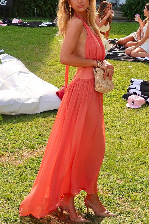 Woman wearing a coral halter neck maxi dress with a flowy skirt and a small straw bag, posing outdoors on a grassy lawn.