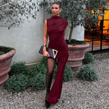 A woman in a burgundy draped dress with an asymmetrical hemline and a thigh-high slit, standing in an outdoor setting with plants and a textured wall.