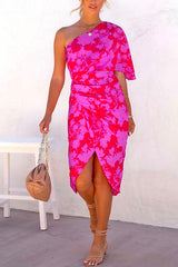 A woman models a one-shoulder pink and red floral print dress with a tulip hem and ruching on the side. She holds a beige clutch bag with a gold chain strap and wears nude strappy heels.