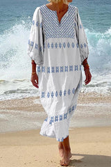 Woman in a white maxi dress with blue embroidery on the neckline and sleeves, standing on a beach with waves in the background.