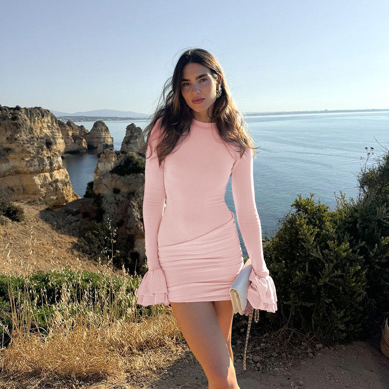 A woman in a light pink ruched mini dress with ruffled sleeves poses with a clutch bag by the sea.