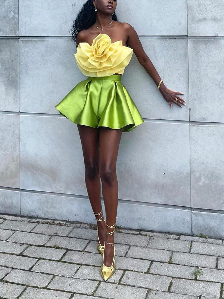 A woman in a yellow floral strapless top and green pleated shorts, with gold heels, standing against a wall.
