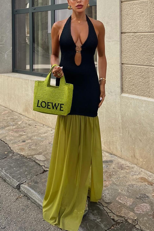 Woman wearing a black halter neck dress with a keyhole cutout and gold hardware detail, paired with a floor-length olive green sheer skirt overlay with a high slit, holding a green woven tote bag, standing on a city street.