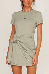 A woman wearing a sage green short-sleeve crew neck dress with a tie waist and ruched detailing.