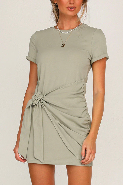 A woman wearing a sage green short-sleeve crew neck dress with a tie waist and ruched detailing.