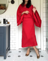 Woman in a red linen dress with kimono sleeves and gold heels, posing in a bathroom.