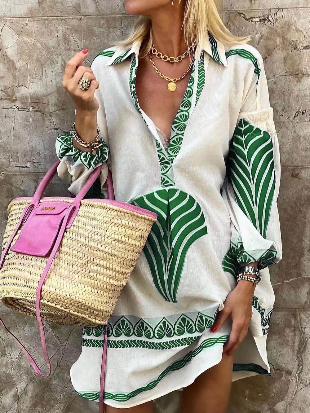 A woman models a white shirt dress with a green leaf print, a straw bag, and gold jewelry.