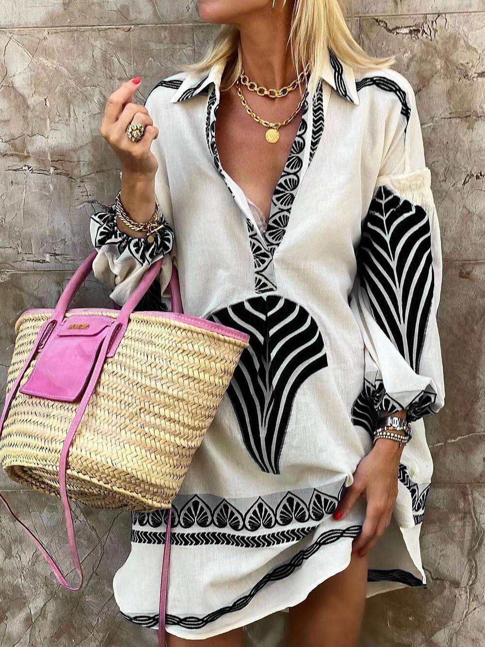 A woman models a white shirt dress with a black leaf print, a straw bag, and gold jewelry.