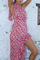 Woman in a pink floral one-shoulder dress with ruffles and a high-low hem, with arms raised against a white wall.