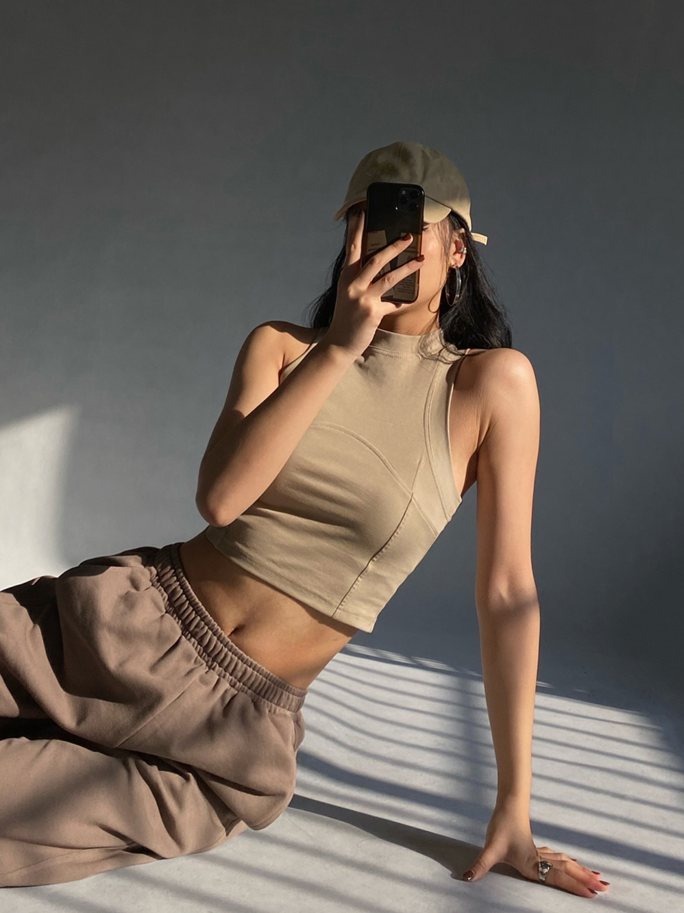 Woman wearing a beige cropped tank top and brown sweatpants, holding a phone to take a selfie.