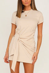 A woman wearing a beige short-sleeve crew neck dress with a tie waist and ruched detailing.