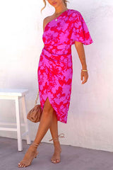 A woman in a one-shoulder pink and red floral print dress poses with her hand on her hip. The dress features a tulip hem and ruching, and she wears nude strappy heels and a delicate necklace.