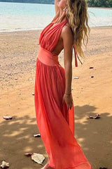Woman wearing a coral halter neck maxi dress with an open back, standing on a sandy beach with the ocean in the background.