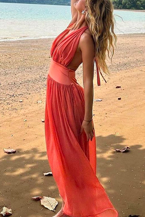 Woman wearing a coral halter neck maxi dress with an open back, standing on a sandy beach with the ocean in the background.