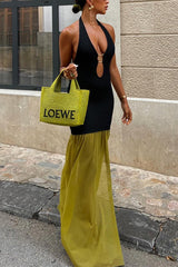 Woman wearing a black halter neck dress with a keyhole cutout and gold hardware detail, paired with a floor-length olive green sheer skirt overlay with a high slit, holding a green woven tote bag, walking on a city street.