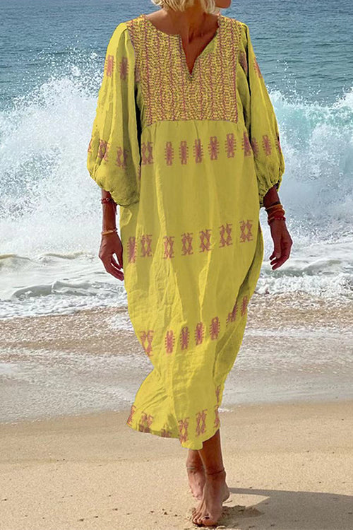 Woman in a yellow maxi dress with pink embroidery on the neckline and sleeves, standing on a beach with waves in the background.