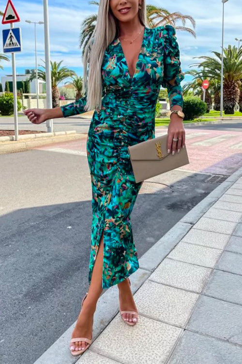 Woman wearing a ruched green floral print v-neck midi dress with puff sleeves and a slit hem, holding a beige clutch and wearing pink heeled sandals.