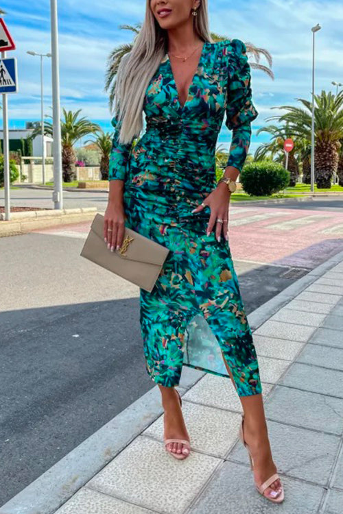 Woman wearing a ruched green floral print v-neck midi dress with puff sleeves and a slit hem, holding a beige clutch and wearing pink heeled sandals.