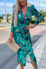 Close-up of a woman wearing a ruched green floral print v-neck midi dress with puff sleeves and a slit hem, holding a beige clutch and wearing pink heeled sandals.