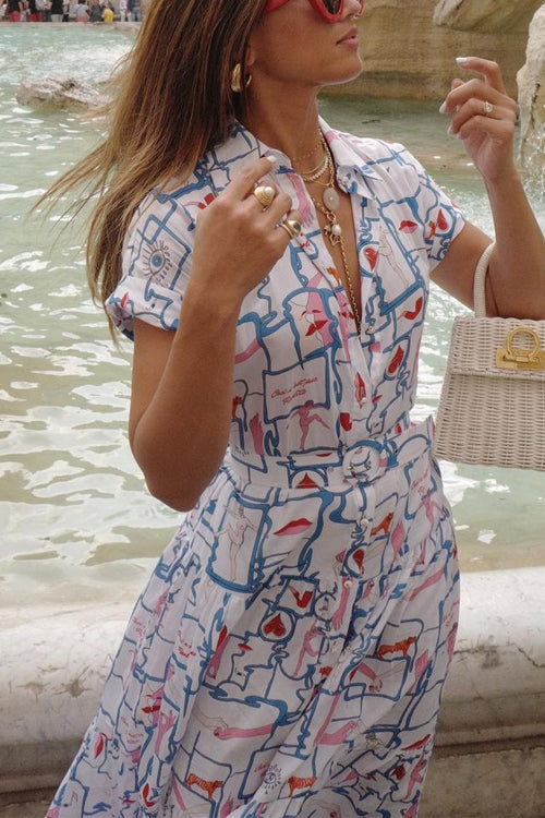 A woman with long blonde hair wearing a printed midi dress and a wicker handbag, with a fountain in the background.