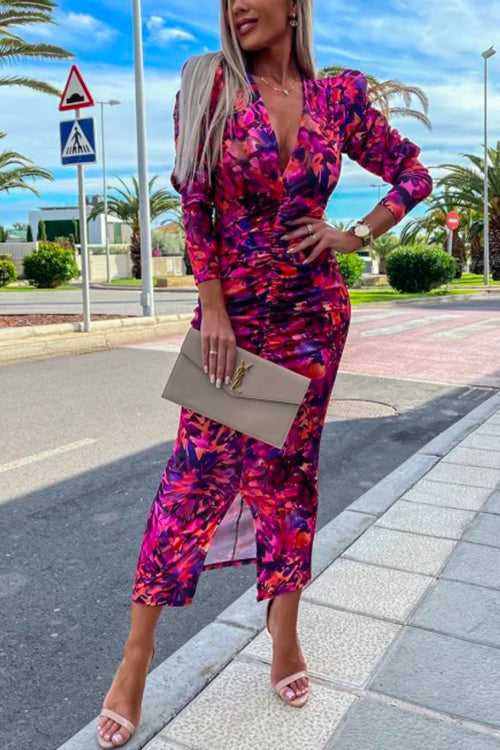 Woman wearing a ruched pink and purple floral print v-neck midi dress with puff sleeves and a slit hem, holding a beige clutch and wearing pink heeled sandals.