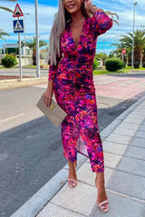 Woman wearing a ruched pink and purple floral print v-neck midi dress with puff sleeves and a slit hem, holding a beige clutch and wearing pink heeled sandals.