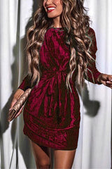Woman in a long-sleeved teal velvet mini dress with a tie waist and mock neck, holding a gold clutch.