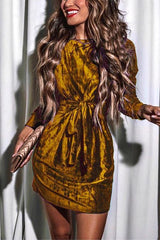 Woman in a long-sleeved emerald green velvet mini dress with a tie waist and mock neck, holding a gold clutch.
