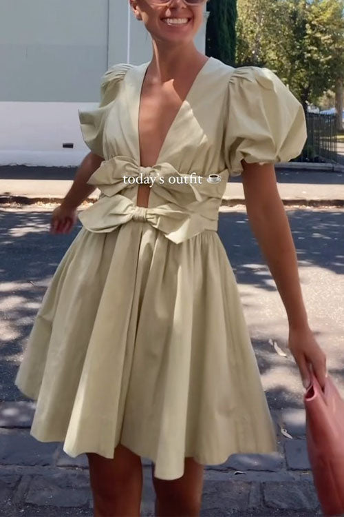 woman wearing a light green puff sleeve dress with a plunging V-neck and bow detail, smiling and holding a pink bag