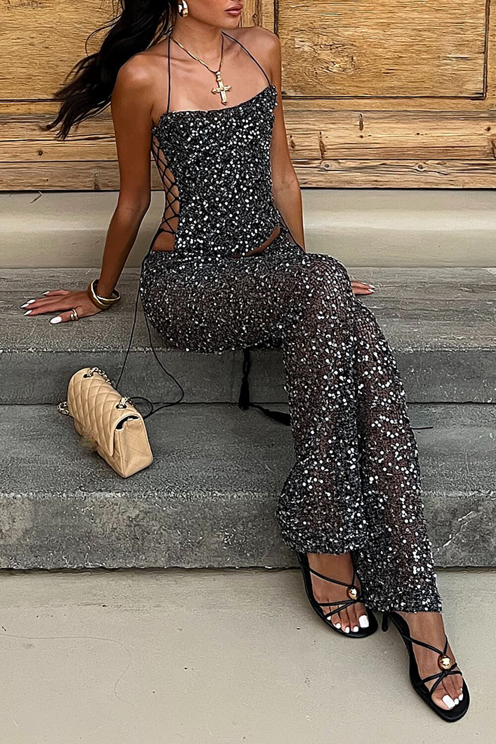 A woman sitting on stone steps wearing a black sequined halter top and matching wide leg pants, with a beige quilted handbag beside her.