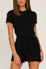 A woman wearing a black short-sleeve crew neck dress with a tie waist and ruched detailing.