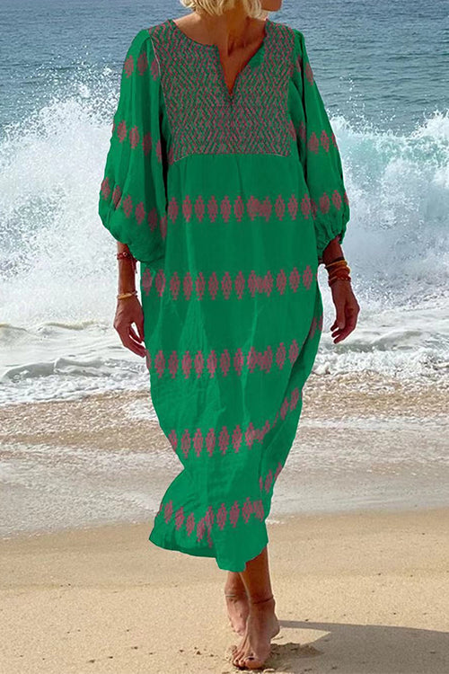 Woman in a green maxi dress with pink embroidery on the neckline and sleeves, standing on a beach with waves in the background.