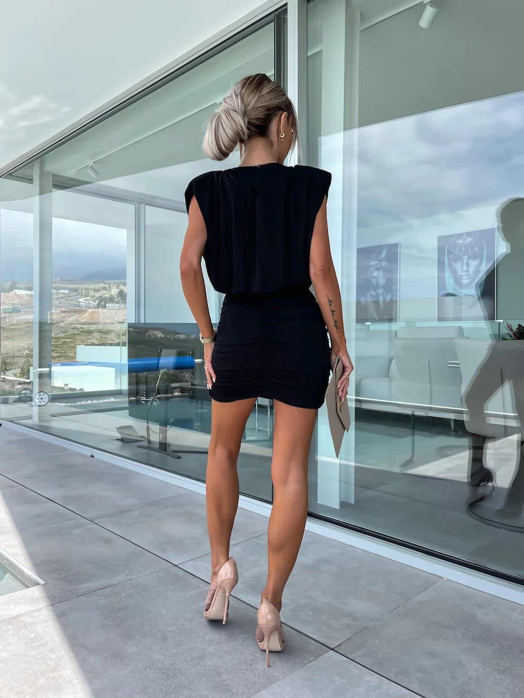 A woman in a black dress with a ruched skirt and padded shoulders, holding a beige clutch bag.