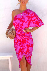 Close-up of a woman wearing a vibrant one-shoulder floral print dress. The dress has a draped effect and a tulip hem. She accessorizes with a beige clutch and gold jewelry.