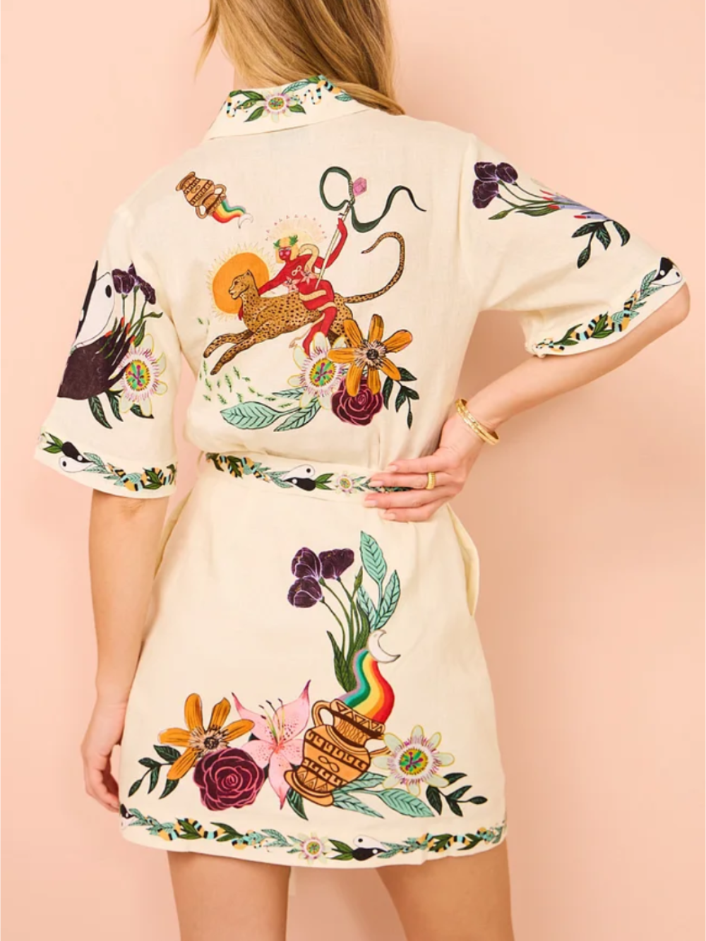 Back view of a woman wearing a cream-colored short-sleeve shirt dress with a colorful floral and animal print, belted at the waist.