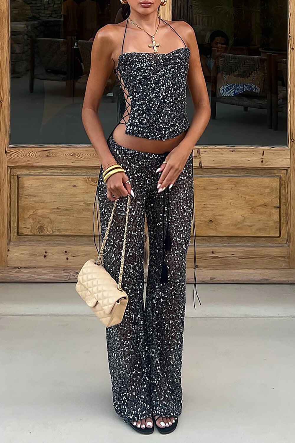 A woman wearing a black sequined halter top and matching wide leg pants, holding a beige quilted handbag.