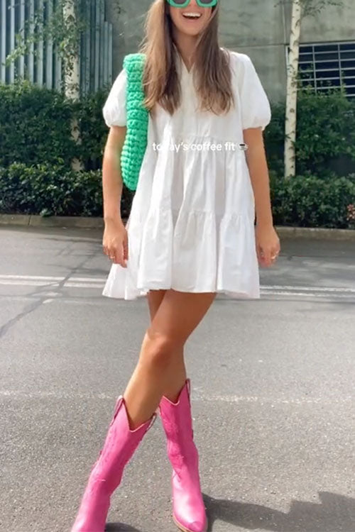 A woman wearing a white tiered dress, bright pink cowboy boots, and a green crochet bag.