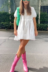 A woman wearing a white tiered dress, bright pink cowboy boots, and a green crochet bag.