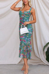 Woman wearing a green floral ruched midi dress with a ruffled mermaid hem and holding a white handbag.