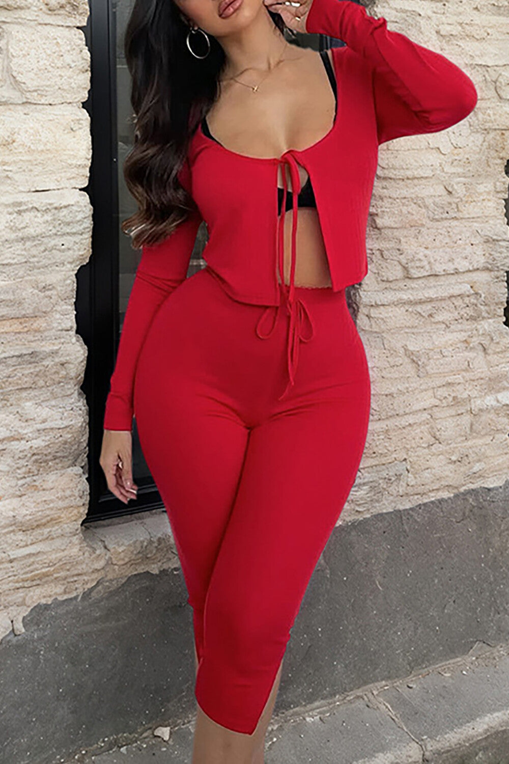 A woman in a red two-piece outfit stands in front of a brick wall. The outfit includes a cropped, tie-front top and matching pants.