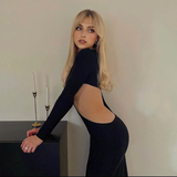 A blonde woman with bangs leans against a dark surface, wearing a long-sleeved black dress with a dramatic side cut-out.