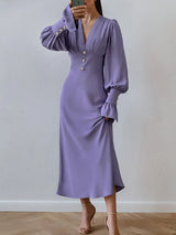 Woman wearing a long-sleeved purple dress with puffed sleeves and a V-neckline, standing and holding the skirt.