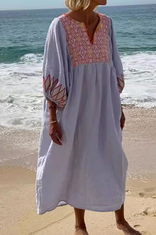 Woman in a light blue maxi dress with pink embroidery on the neckline and sleeves, standing on a beach with waves in the background.