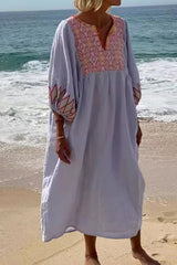 Woman in a light blue maxi dress with pink embroidery on the neckline and sleeves, standing on a beach with waves in the background.