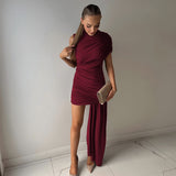 A woman in a burgundy draped dress with an asymmetrical hemline and a thigh-high slit, holding a clutch and looking down.
