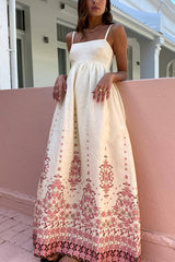 A woman leaning against a pink wall wearing a cream maxi dress with spaghetti straps and a pink floral print.
