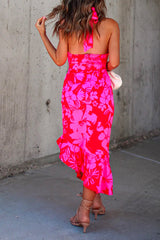 Back view of a woman wearing a bright pink and red floral print halter neck midi dress with a ruffled asymmetrical hem, holding her hair and a white clutch, standing in front of a concrete wall.